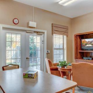 A cozy living room with a table, chairs, and a television near bright windows and a glass door.