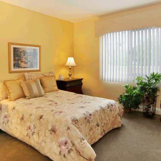 A cozy bedroom with a warm color scheme, featuring a bed with floral bedding, a bedside lamp, and a window with blinds.