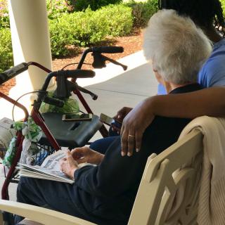 An elderly person sits on a bench outdoors with a caregiver by their side, showing companionship and support.