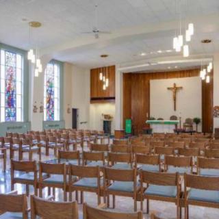 An empty church interior featuring wooden chairs facing an altar with a cross, illuminated by natural light through large windows.