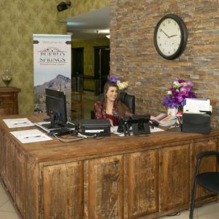 A receptionist sits at a wooden front desk in a hotel lobby, with decor including a clock and flower arrangements.