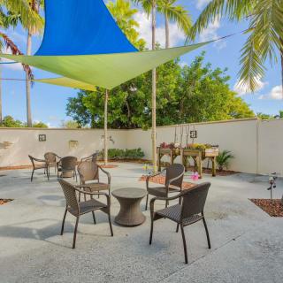 A sunny outdoor patio area with chairs and tables under a colorful shade sail, surrounded by palm trees.