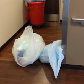 Two bags of waste positioned near a door in a hallway with a red trash bin in the background.