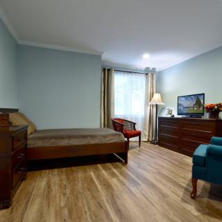 A cozy bedroom with wood flooring, featuring a bed, dresser, chairs, and a television.
