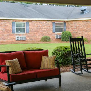 A cozy outdoor seating area featuring a red couch and a rocking chair, with a lush green lawn and a brick house in the background.