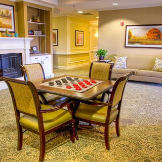 A cozy living room featuring a checkers board set up on a table surrounded by chairs, with a fireplace and artwork in the background.