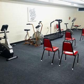 A small gym room with exercise equipment including stationary bikes and a treadmill, alongside a few red chairs.
