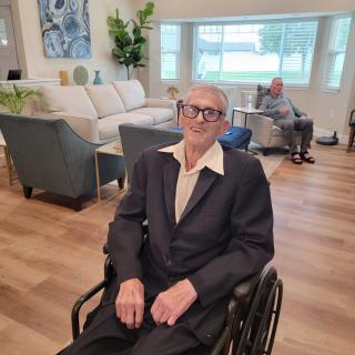 An elderly man wearing glasses is sitting in a wheelchair in a living room.