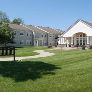 A peaceful residential building surrounded by a green lawn and trees with a bench along a curved pathway.