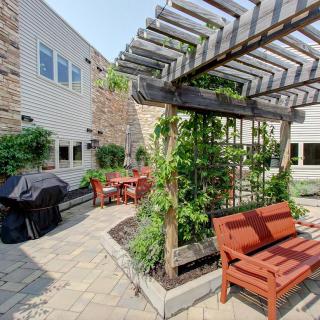 A charming outdoor patio with a pergola, featuring wooden benches and chairs surrounded by lush greenery.