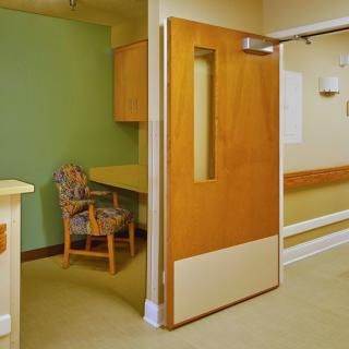 A hospital hallway with an open door leading to a room equipped with a chair and desk.