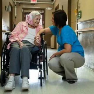 A caregiver kneels to speak with an elderly woman in a wheelchair in a hallway.