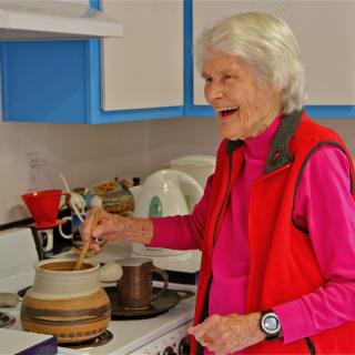An elderly woman smiles while cooking in the kitchen.