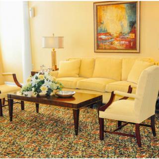 A cozy living room with a yellow sofa, two armchairs, and a wooden coffee table adorned with flowers, complemented by a patterned rug and a lamp next to abstract wall art.