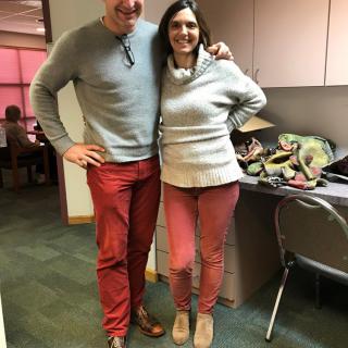 Two colleagues wearing matching red pants and grey sweaters pose together in an office.