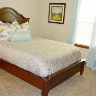 A cozy bedroom featuring a neatly made bed with decorative pillows, soft carpeting, and a framed artwork on the wall.