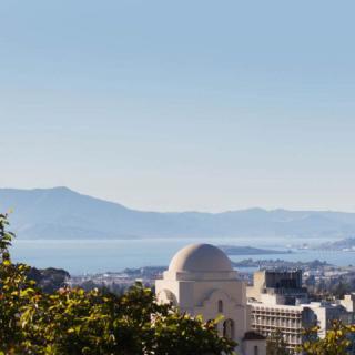 A scenic view of a city with a prominent dome-shaped building, overlooking the ocean and distant mountains.