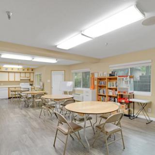 A spacious classroom with round tables, folding chairs, bookshelves, and bright overhead lighting.