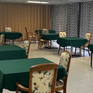 A dining room with several tables covered in green tablecloths and floral-patterned chairs, set against a backdrop of curtains.