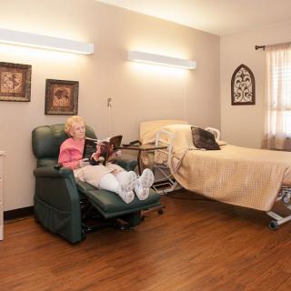 An elderly person sitting in a recliner in a nursing home room with a bed and framed pictures on the walls.