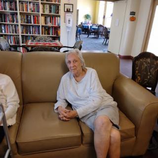 An elderly woman relaxing on a sofa in a communal space with bookshelves in the background.