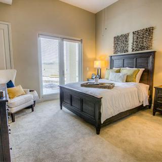 A cozy, well-decorated bedroom featuring a large bed with accent pillows, a lamp on the bedside table, and natural light streaming through a window.