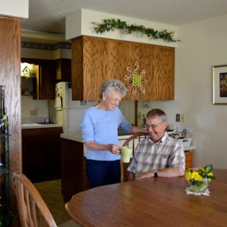 An elderly woman assists a man seated at the dining table in a cozy kitchen setting.
