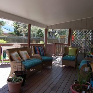 A cozy porch with wicker furniture and colorful cushions, surrounded by greenery in natural sunlight.