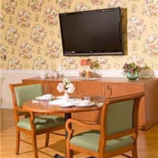 A cozy dining area featuring a round table with chairs, a cabinet, and a wall-mounted television against floral wallpaper.