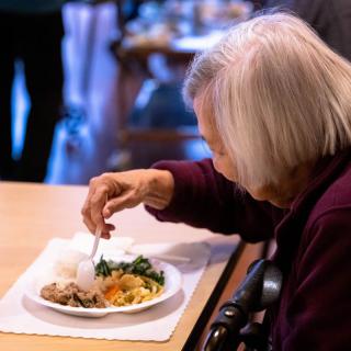 An elderly woman is sitting at a table, enjoying her meal in an indoor setting.