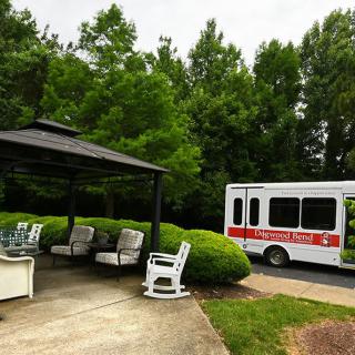 A quiet outdoor seating area next to a white bus with a red stripe, surrounded by lush greenery and trees.