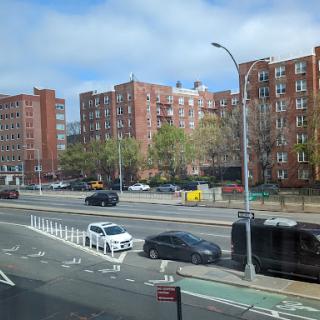 A view of a city street lined with residential buildings and parked cars under a partly cloudy sky.