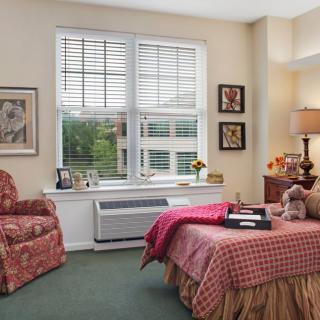 A cozy bedroom with a large window, an armchair, and a neatly made bed with a red bedspread.