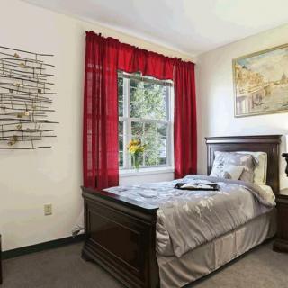 A cozy bedroom featuring a single bed with gray bedding, red curtains framing a window, and decorative wall art.