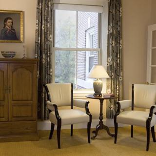 A cozy living room corner featuring elegant chairs, a lamp on a side table, and a large window with curtains.