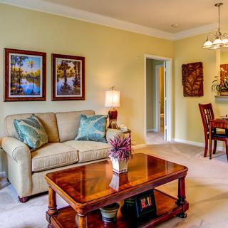 A cozy living room featuring a comfortable sofa, wooden coffee table, and decorative artwork on the walls. The image also shows an adjoining dining area with a chandelier.
