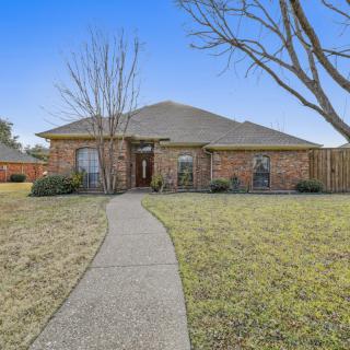 A single-story brick house with a pathway leading to the entrance, surrounded by a lawn and trees.