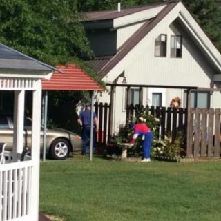 A small house with a garden, a parked car, and people working in the yard.