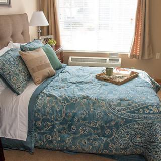 A cozy bedroom featuring a neatly made bed with patterned blue quilt and pillows, a tray on top, and natural light coming through the window.