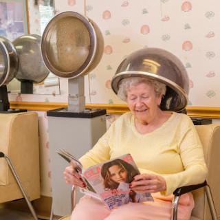 An elderly woman enjoys reading a magazine while undergoing a beauty treatment at a hair salon.