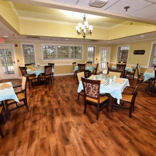 A cozy dining room with neatly arranged tables and chairs, featuring a wooden floor and chandelier.