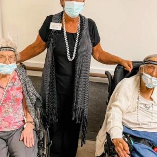 A caretaker standing between two elderly individuals seated in wheelchairs, all wearing masks.