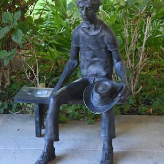 A bronze sculpture of a seated man holding a hat, surrounded by lush greenery.