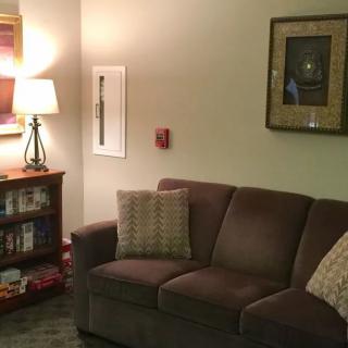 A cozy living room featuring a comfortable brown sofa, illuminated by a lamp on the side table. The room is decorated with framed artwork on the walls and books neatly arranged in a bookshelf.