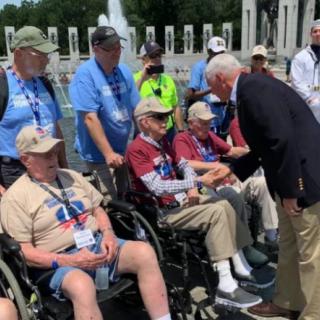 An outdoor gathering where a group of veterans, some in wheelchairs, are being greeted with a handshake.