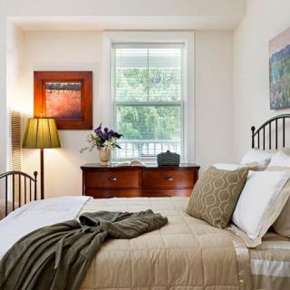 A cozy bedroom featuring a neatly made bed with pillows and a blanket, a window letting in natural light, and a dresser topped with flowers and a lamp.