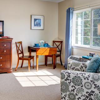 A cozy living room with a table set, chairs, a chest of drawers, and sunlight streaming through the window.