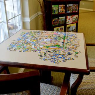 A table with an incomplete jigsaw puzzle near a window, surrounded by chairs and a bookshelf in the background.