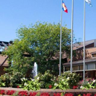 A sunny day view of a building with flags, surrounded by a garden with flowers and a fountain.