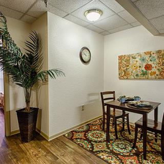 A cozy dining room with a small wooden table set, vibrant rug, decorative wall art, and a potted plant.